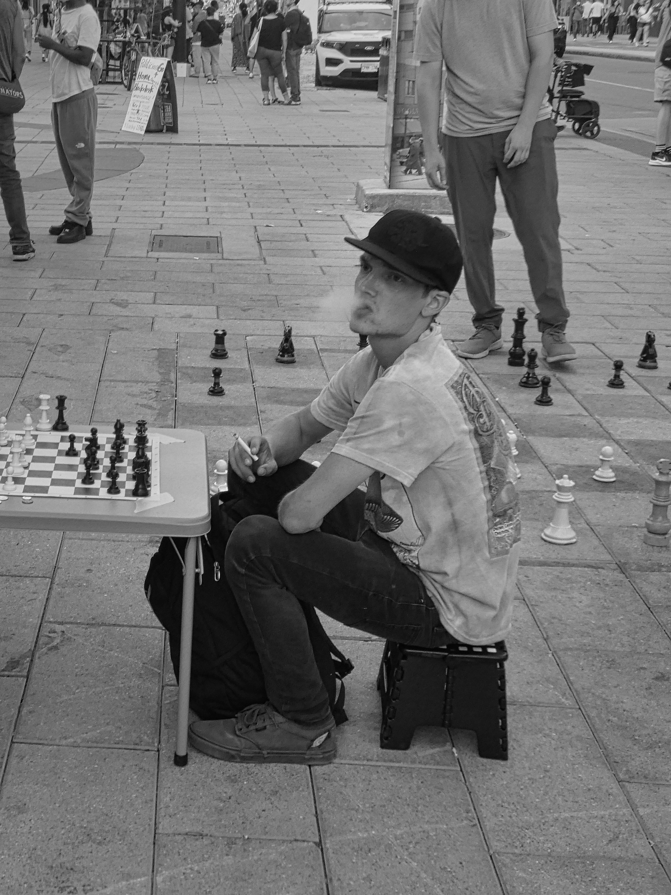 a young man blow out cigarette smoke as he plays chess on a city sidewalk