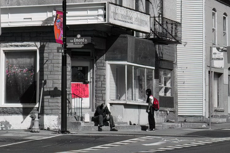 a young woman speaks to a man sitting on the stoop of a vacant store/
