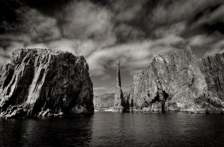 A rugged spire rises from the sea on a rugged coastline.